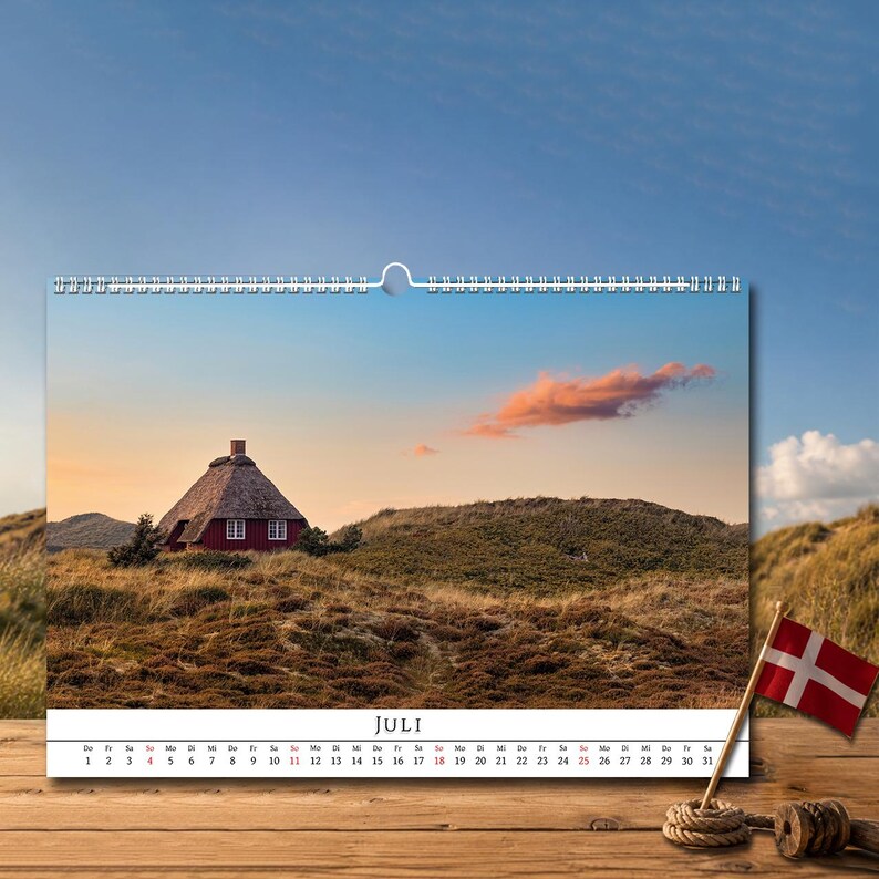 Op de afbeelding: Een wandkalender voor de maand juli met een foto van een rood huis met een rieten dak in een grasachtig duinlandschap onder een zonsondergang. De kalender bevat een datumsraster en een kleine Deense vlag op een houten stokje.