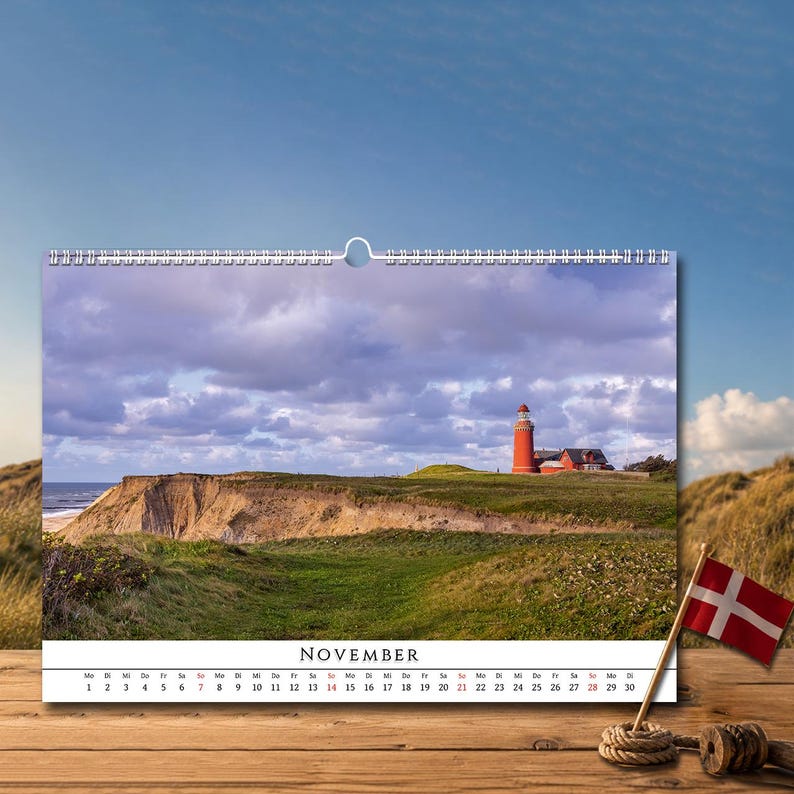 Op de afbeelding: Een wandkalender voor november met een foto van een rode vuurtoren op een grasrijke klif die uitkijkt over de zee onder een bewolkte lucht. De kalender bevat een raster met datums en de maandnaam, geplaatst op een houten ondergrond.