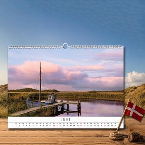 Op de afbeelding: Een wandkalenderpagina voor de maand juni met een foto van een blauwe boot aangemeerd aan een houten steiger in een rustig water. De sc&egrave;ne bevat hoog gras, een zachtroze en blauwe lucht en de tekst JUNI onderaan met de bijbehorende data.