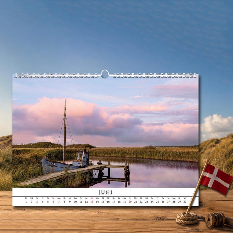 Op de afbeelding: Een wandkalenderpagina voor de maand juni met een foto van een blauwe boot aangemeerd aan een houten steiger in een rustig water. De sc&egrave;ne bevat hoog gras, een zachtroze en blauwe lucht en de tekst JUNI onderaan met de bijbehorende data.