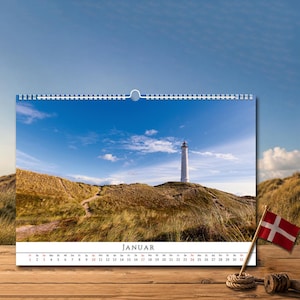 Op de afbeelding: Een wandkalender met een foto van een witte vuurtoren op een grasduin onder een blauwe lucht. De pagina toont de maand JANUAR met een datumrooster onder de afbeelding. Een kleine Deense vlag aan een houten stok staat aan de rechterkant.