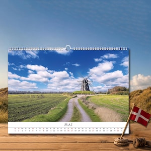 Op de afbeelding: Een wandkalender met een landschapsfoto van een traditionele windmolen in een groen veld onder een blauwe lucht met witte wolken. De kalenderpagina toont de maand MAI met een raster van data van 1 tot 31 in zwarte en rode tekst.