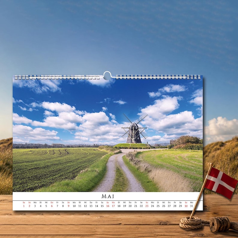 Op de afbeelding: Een wandkalender met een landschapsfoto van een traditionele windmolen in een groen veld onder een blauwe lucht met witte wolken. De kalenderpagina toont de maand MAI met een raster van data van 1 tot 31 in zwarte en rode tekst.