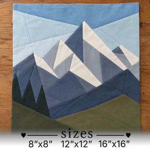 May include: A fabric quilt block featuring a geometric mountain landscape in shades of blue, white, and olive green. The design includes angular peaks and dark green trees. Text at the bottom lists available sizes as 8"x8", 12"x12", and 16"x16".