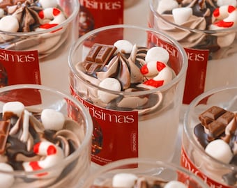 Pode incluir: Velas em copos transparentes, com aspeto de sobremesa. Apresentam espirais de chocolate, marshmallows e pedaços de barras de chocolate. Etiquetas vermelhas com a palavra "Christmas" são visíveis.