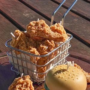 May include: A selection of food-shaped candles, featuring fried chicken pieces in a metal basket and a hamburger. The fried chicken candles are golden brown, and the hamburger candle has a sesame seed bun.