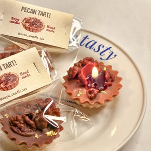 Könnte beinhalten: Pekannuss-Törtchen-Kerzen mit einer brennenden Kerze, Walnüssen und goldenen Blattakzenten. Die Kerzen sind in Klarsichtfolie verpackt und stehen auf einem weißen Teller mit dem Wort "tasty".