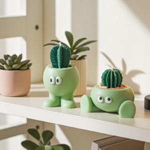 Könnte beinhalten: Zwei grüne Kerzen in Kaktusform mit weißen Augen und einem grünen Kaktusdesign. Eine Kerze steht aufrecht, die andere liegt. Eine kleine Sukkulentenpflanze in einem rosa Topf ist ebenfalls zu sehen.