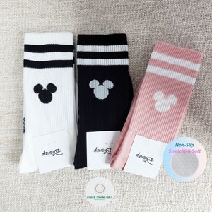 Könnte beinhalten: Drei Paar Socken mit einem Mickey Mouse-Motiv. Die Socken sind weiß mit schwarzen Streifen, schwarz mit weißen Streifen und rosa mit weißen Streifen. Alle Socken sind mit dem Disney-Logo versehen.