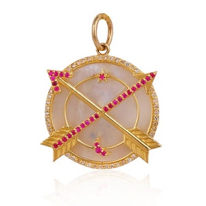 Może przedstawiać: Złoty naszyjnik z zawieszką w kształcie okrągłego charmsa z perłowej muszli, na którym znajdują się dwie skrzyżowane strzały z czerwonymi rubinowymi akcentami i diamentową aureolą. Charms ma również wzór półksiężyca i gwiazdy.