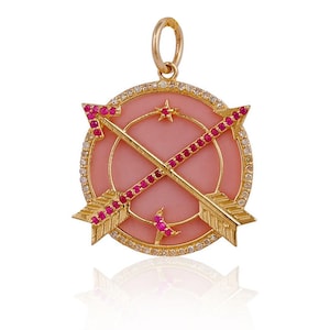 Może przedstawiać: Złoty wisiorek z różowym kamieniem i dwoma skrzyżowanymi strzałami ozdobionymi czerwonymi kamieniami szlachetnymi. Wisiorek jest otoczony kółkiem z małych diamentów.