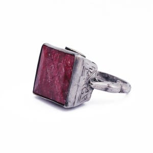 Może przedstawiać: Srebrny pierścionek z kwadratowym, czerwonym kamieniem szlachetnym osadzonym w środku. Obrączka pierścionka jest ozdobiona misternymi wzorami.