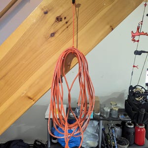 May include: Orange extension cord hanging from a black hook. The cord is coiled and suspended against a wooden staircase. The background includes a shelf with various items, including jars and sports equipment.