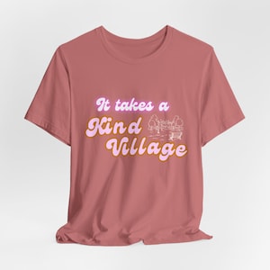 May include: A dusty rose t-shirt with the phrase "It takes a Kind Village" in a retro font. The text is outlined in brown and has a light purple glow. A small illustration of a park scene is next to the text.