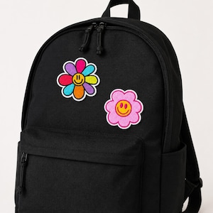 Peut inclure: Un sac à dos noir orné de deux écussons en forme de fleur brodés sur la poche avant. L'un est une fleur multicolore aux pétales roses, violets, bleus, jaunes et orange, l'autre est une fleur rose unie. Les deux présentent des visages souriants jaunes.