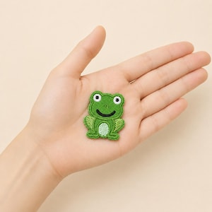 May include: An embroidered iron-on patch featuring a small green frog with a light green belly, black stitched eyes, and a curved black mouth. The frog is depicted in a seated position with its front legs resting on its sides and back legs tucked.