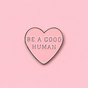 May include: A heart-shaped enamel pin with a light pink surface and a thin silver-toned metal border. The text BE A GOOD HUMAN is printed in black capital letters across the centre of the heart, following the shape of the pin's design.