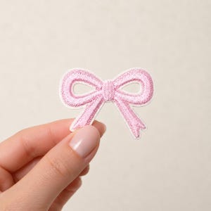 Op de afbeelding: Een lichtroze geborduurde strikpatch met een gestikte textuur en een gedefinieerde knoop in het midden. Het ontwerp bestaat uit twee symmetrische lussen en twee korte, schuine lintuiteinden die naar beneden wijzen, gemaakt met dicht stikwerk.