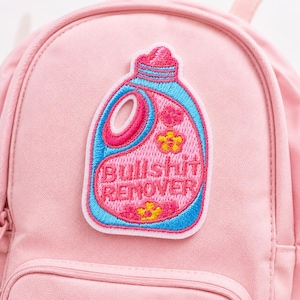 May include: A light pink backpack featuring an embroidered patch of a detergent bottle in shades of blue, pink, and yellow. The patch displays the text "BULLSHIT REMOVER" in bold, capitalized letters across the center of the bottle design.