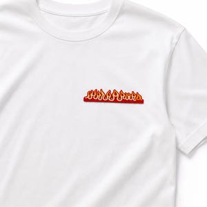 Peut inclure: Un t-shirt blanc à manches courtes avec un petit écusson brodé horizontal sur la poitrine gauche. L'écusson présente un motif de flammes stylisé aux couleurs vives rouge, orange et jaune, souligné par une bordure rouge marquée.
