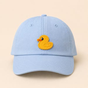 May include: A light blue baseball cap featuring an embroidered yellow rubber duck patch on the front center. The cap has a structured crown, a curved brim, and matching blue stitching details throughout the fabric construction.