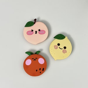 May include: Three silicone charms shaped like fruits with cartoon faces. One is a light pink peach with a green leaf, one is a yellow lemon with a green leaf and dotted texture, and one is a round orange fruit with a green leaf and pink cheeks.