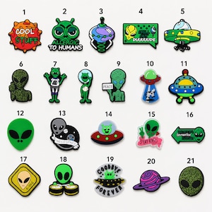 May include: A collection of twenty-one colourful silicone shoe charms featuring various alien, UFO, and space-themed designs. Each charm displays unique graphics including text like COOL STUFF, ALLERGIC TO HUMANS, CHILL OUT DUUUUUDE, PEACE, STAY RAD, and GOODBYE FOREVER.