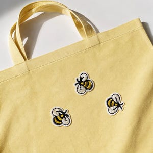Könnte beinhalten: Eine hellgelbe Tragetasche aus Segeltuch mit drei aufgestickten Bienen-Applikationen. Jede Biene ist mit schwarzem, gelbem und weißem Garn dargestellt und zeigt detaillierte Flügel sowie gestreifte Körper. Die Tasche hat zwei passende gelbe Griffe.
