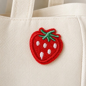 May include: A cream-colored canvas tote bag featuring a centered, embroidered strawberry patch. The strawberry is bright red with a green stem and small white embroidered seeds, showcasing a textured, stitched finish on the fabric surface.
