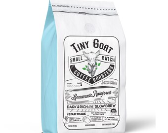 Puede incluir: Una bolsa blanca de café molido Tiny Goat Small Batch Coffee Roasters Baaamaste Rainforest Dark & Rich for Slow Brew. La bolsa presenta una cabra de dibujos animados y el texto "Clean Roasted - No VOCs or Emissions". La bolsa es de 454 g y está certificada por Fair Trade.