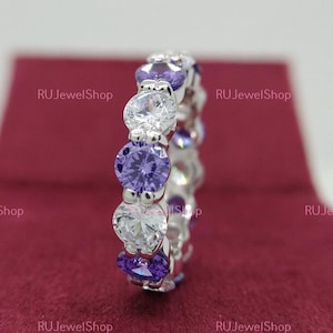 May include: A silver band ring with alternating clear and purple round stones. The ring is on a red velvet background.
