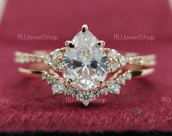 May include: A pear-shaped diamond engagement ring set in rose gold with a delicate band and a halo of smaller diamonds. The ring is displayed on a red velvet box.