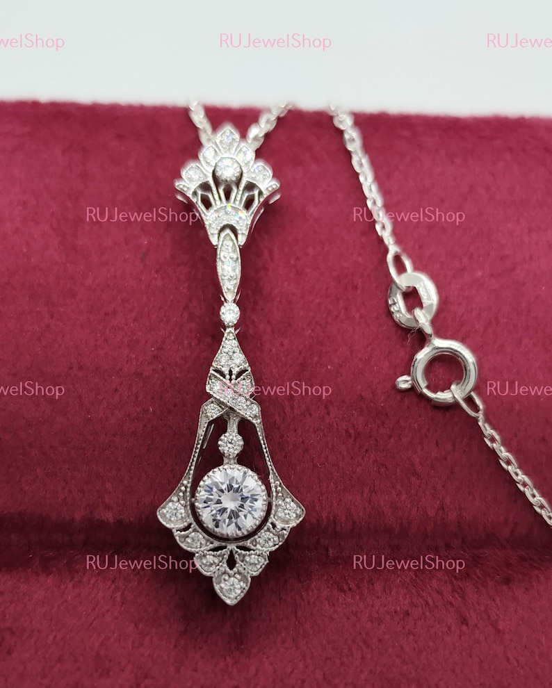 May include: A silver necklace with a pendant featuring a large round clear stone surrounded by smaller stones. The pendant is in an art deco style with intricate details. The necklace is on a red velvet background.