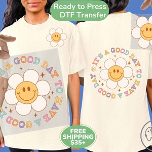 May include: A white t-shirt with a colorful graphic design that says "It's a good day to have a good day". The graphic features a smiling flower with a yellow face and white petals. The design is on a clear transfer sheet.