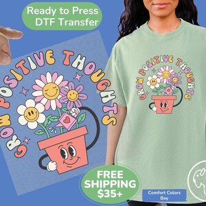 May include: A light green t-shirt with a graphic of a flower pot with smiling eyes and a watering can. The flowers in the pot have smiling faces and the text around the pot reads "Grow Positive Thoughts".