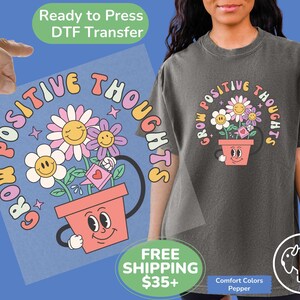 May include: A light gray t-shirt with a graphic design of a flower pot with a smiling face. The pot is filled with flowers and the text "Grow Positive Thoughts" is written in a circle around the pot.