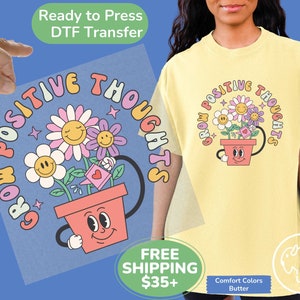 May include: A yellow t-shirt with a graphic of a flower pot with a smiling face and three flowers growing out of it. The text "Grow Positive Thoughts" is written in a rainbow-colored arc around the pot.