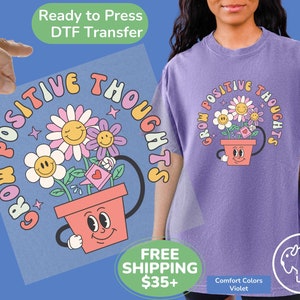 May include: A purple t-shirt with a graphic of a flower pot with smiling flowers and the text "Grow Positive Thoughts".
