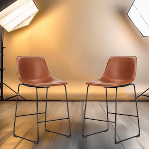 May include: Two brown leather bar stools with black metal frames. The stools are set against a backdrop of a brick wall and a light brown backdrop.