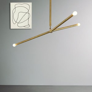 May include: A brass three-arm chandelier with three light bulbs. The chandelier is hanging from the ceiling in a room with a white wall and a wooden floor. The text "Lampize Studio" and "20% OFF" is visible in the image.