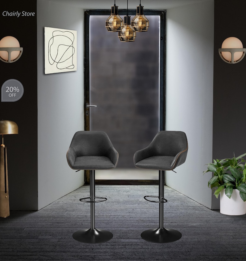 Puede incluir: Dos taburetes de bar giratorios tapizados en gris con bases y reposapi&eacute;s de metal negro. Los taburetes est&aacute;n en una habitaci&oacute;n con una puerta, una l&aacute;mpara y una pared con una pintura abstracta en blanco y negro.