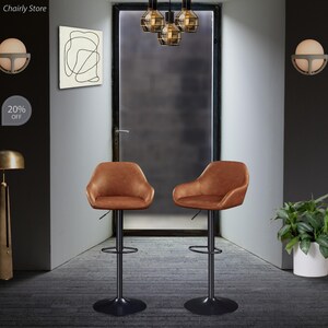 Puede incluir: Dos taburetes de bar giratorios de cuero marr&oacute;n con marcos y reposapi&eacute;s de metal negro. Los taburetes est&aacute;n en una habitaci&oacute;n con una puerta, una l&aacute;mpara y una pared con una pintura abstracta en blanco y negro.