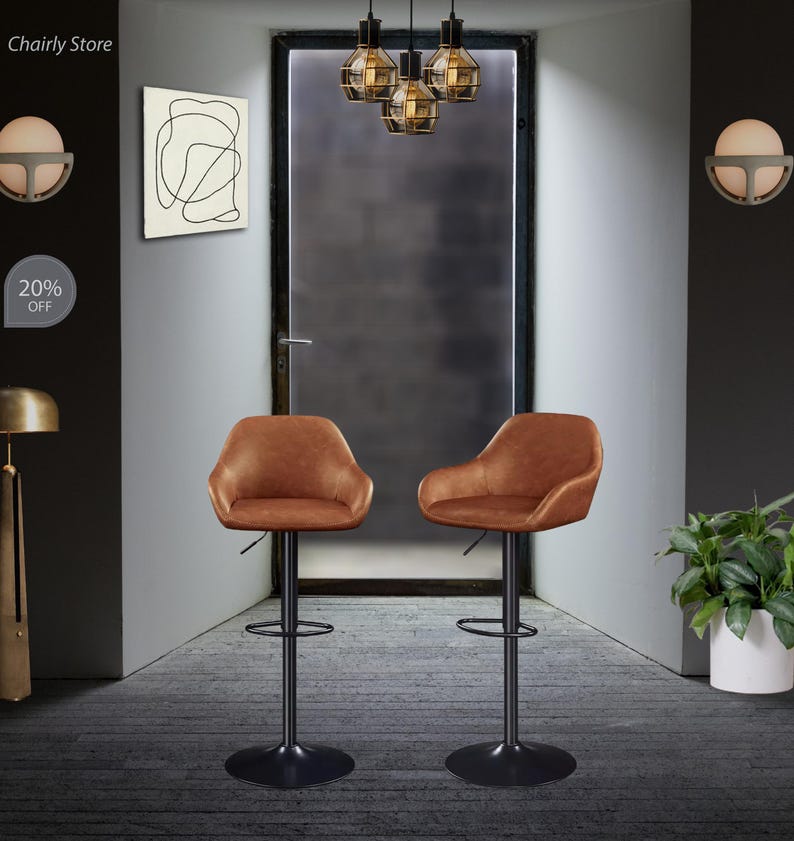 Puede incluir: Dos taburetes de bar giratorios de cuero marr&oacute;n con marcos y reposapi&eacute;s de metal negro. Los taburetes est&aacute;n en una habitaci&oacute;n con una puerta, una l&aacute;mpara y una pared con una pintura abstracta en blanco y negro.