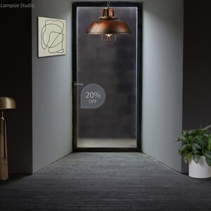 May include: A rusty brown metal pendant light with a cage-like shade hangs from the ceiling in a room with a gray wall and a door. The door has a frosted glass panel with a white sticker that says "20% OFF".
