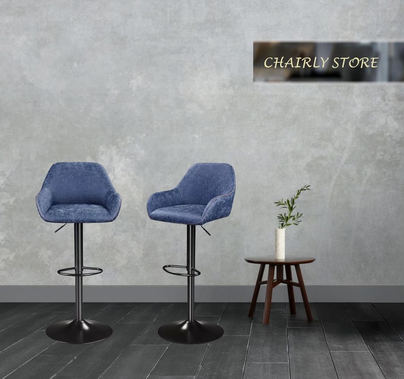 Puede incluir: Dos taburetes de bar giratorios tapizados en azul con bases de metal negro. Los taburetes tienen un respaldo ligeramente curvo y apoyabrazos. Los taburetes est&aacute;n colocados contra una pared gris con un letrero que dice "Chairly Store".