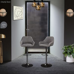 Puede incluir: Dos taburetes de bar tapizados en gris con bases y reposapi&eacute;s de metal negro. Los taburetes est&aacute;n en una habitaci&oacute;n con una puerta y una l&aacute;mpara colgando del techo.