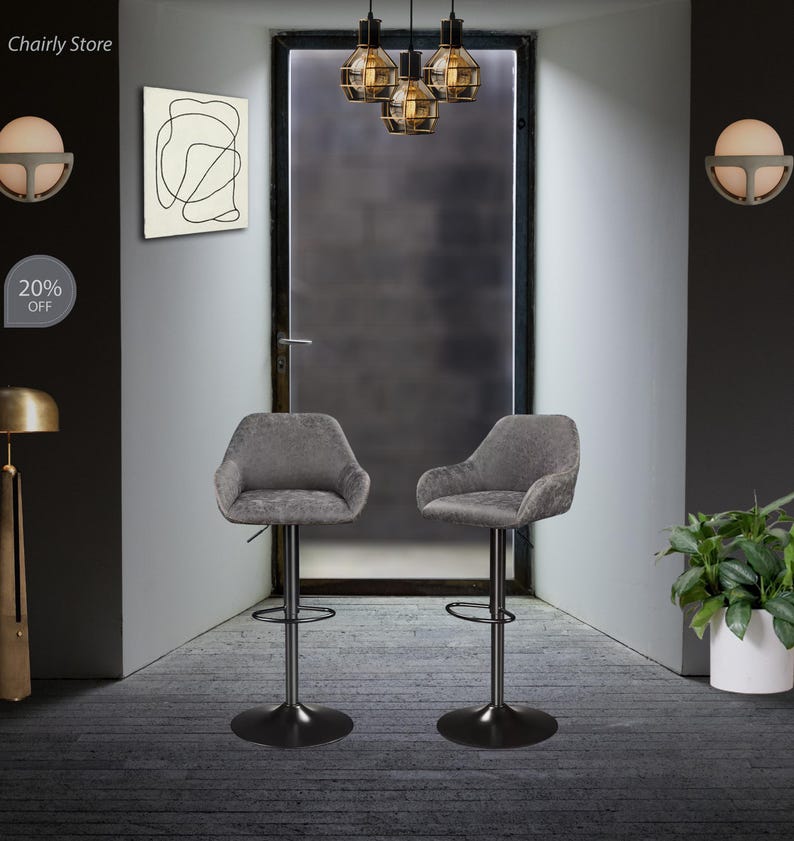 Puede incluir: Dos taburetes de bar tapizados en gris con bases y reposapi&eacute;s de metal negro. Los taburetes est&aacute;n en una habitaci&oacute;n con una puerta y una l&aacute;mpara colgando del techo.
