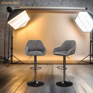 Puede incluir: Dos taburetes de bar giratorios tapizados en gris con bases de metal negro. Los taburetes est&aacute;n instalados en un estudio con fondo e iluminaci&oacute;n.
