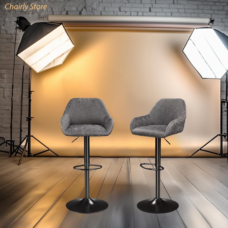 Puede incluir: Dos taburetes de bar giratorios tapizados en gris con bases de metal negro. Los taburetes est&aacute;n instalados en un estudio con fondo e iluminaci&oacute;n.