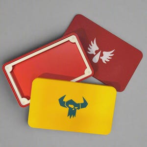 May include: A rectangular yellow storage tin featuring a dark teal skull icon with horns, alongside a red lid with a white winged emblem and an open red container with a white interior frame. These metal boxes have rounded corners and flat surfaces.